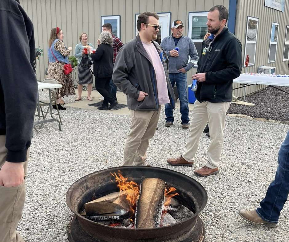 People are gathered outdoors on a gravel surface, chatting around a small fire pit. The mood appears relaxed and social.