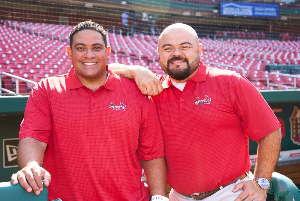 two LatinX men in red shirts stand in sunshine in front of rows of green stadium seats