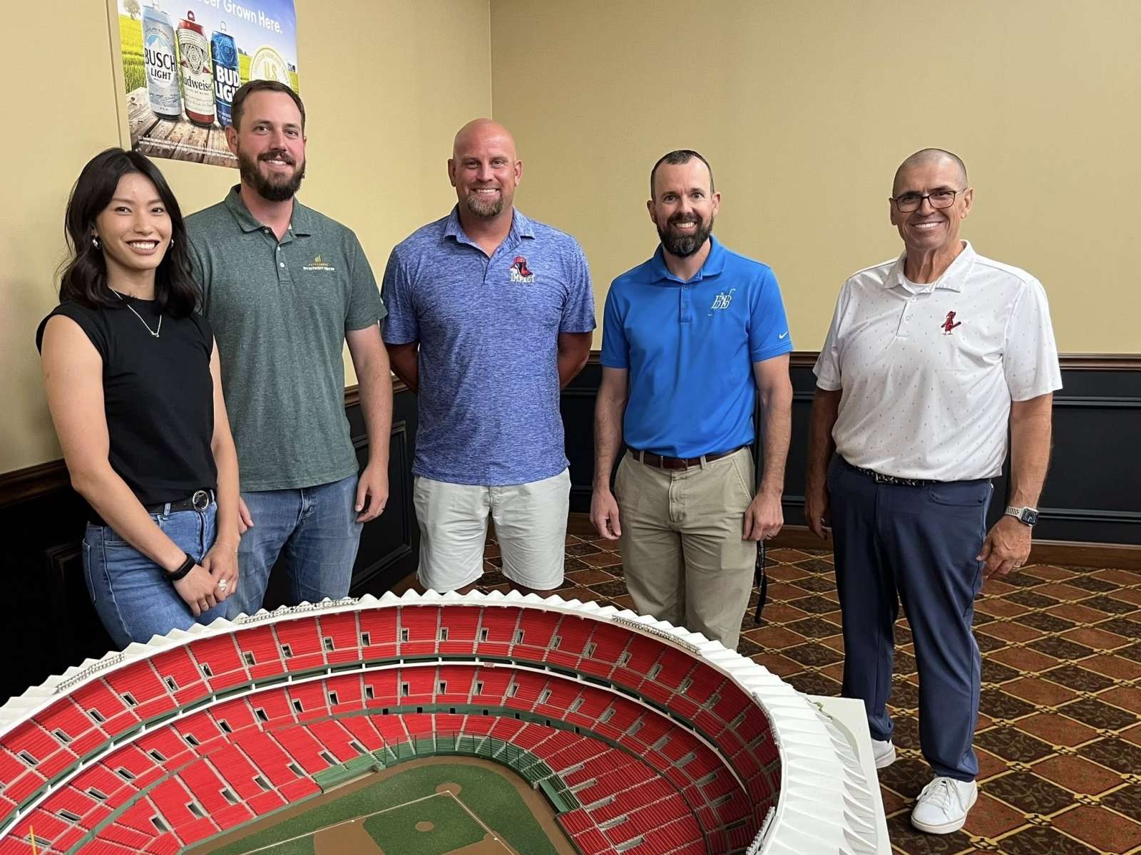 four men in polo shirts and one woman and stand smiling indoors by a scale model of a baseball park.
