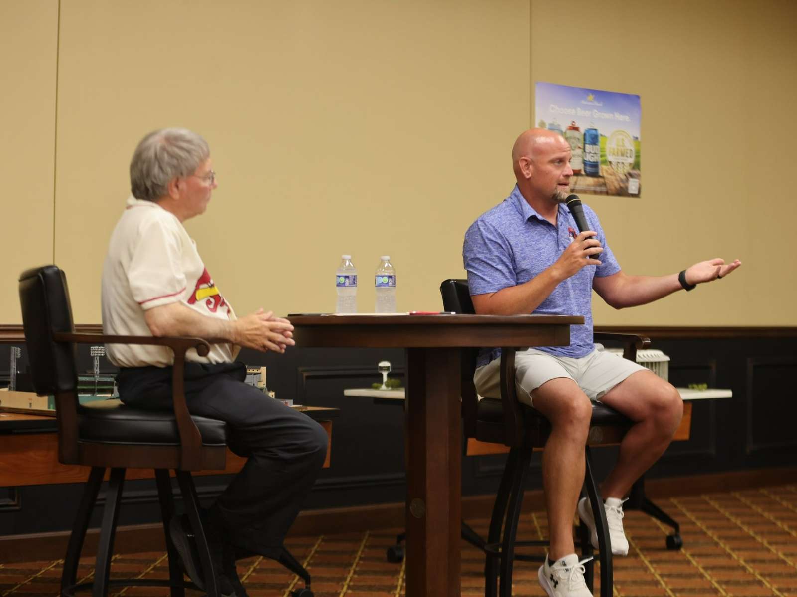 bald athletic man speaks into handheld microphone, answering a question posed by a gray haired man wearing St Louis Cardinals baseball jersey.
