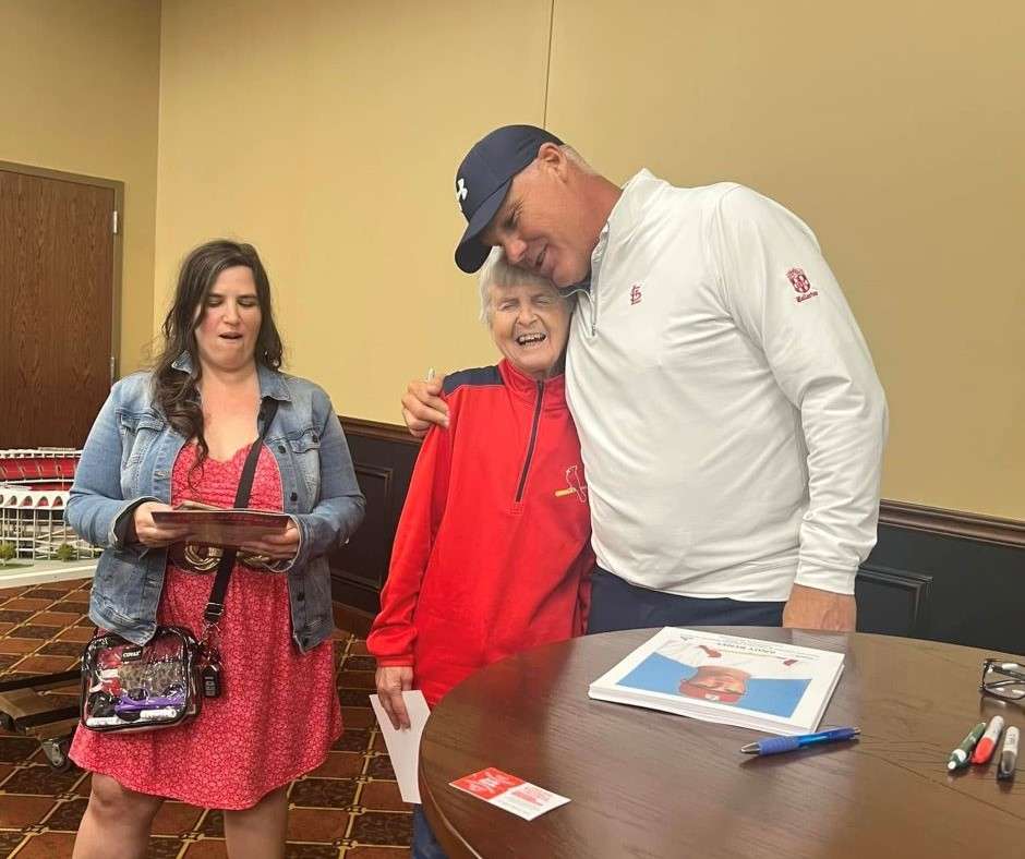 A man in a white shirt and cap hugs an elderly woman in red, both smiling warmly. A woman in a denim jacket and red dress stands nearby, holding papers. They are in a room with a wooden table and markers, conveying a warm and cheerful atmosphere.