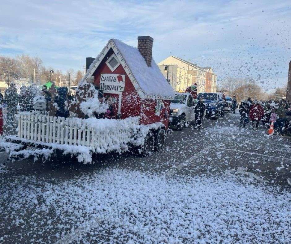 A festive parade features a red Santa's Workshop float covered in artificial snow and foam blowing about, with people and vehicles in the background. The atmosphere is joyful and wintery.