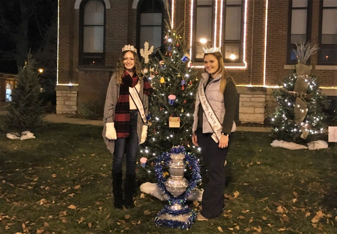 two ladies dressed in warm clothes standing on either side of a 6 foot green evergreen tree decorated with lights and ornaments