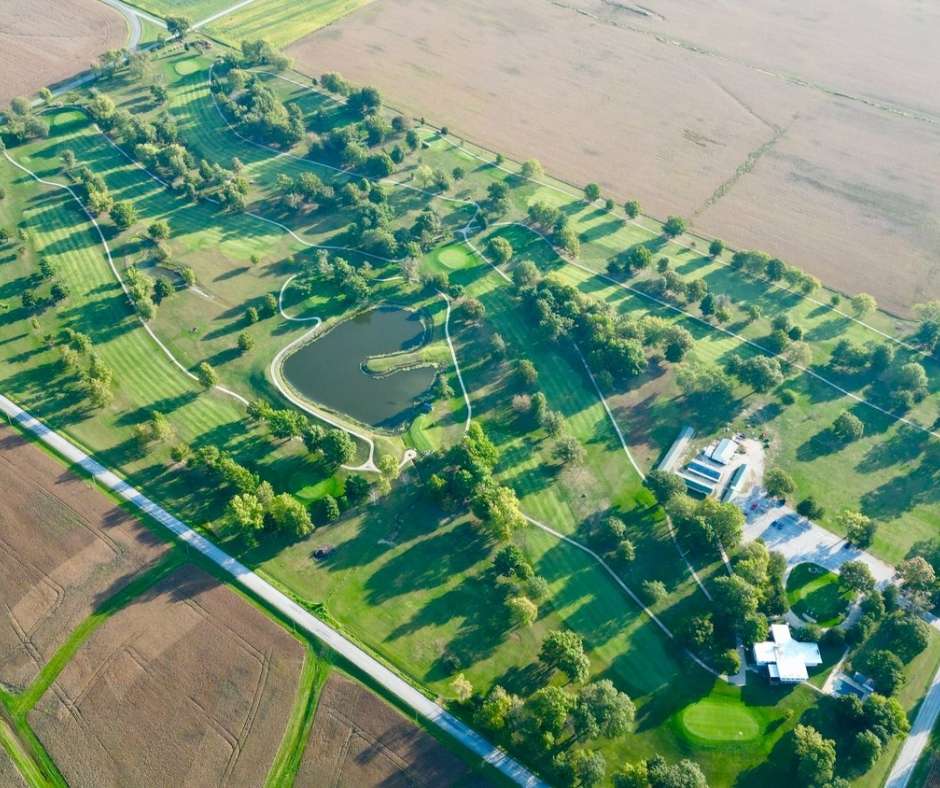 Aerial view of a golf course surrounded by farmland. The landscape features green fairways, a pond, numerous trees, and adjacent fields under clear skies.