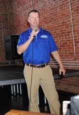 younger middle aged man with short hair wearing royal blue polo shirt and tan trousers stands and holds microphone with cord in front of a brick wall and black stage.