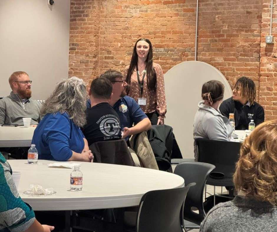 A woman standing and speaking in a room with a brick wall, surrounded by seated people at round tables. The atmosphere is lively and engaging.