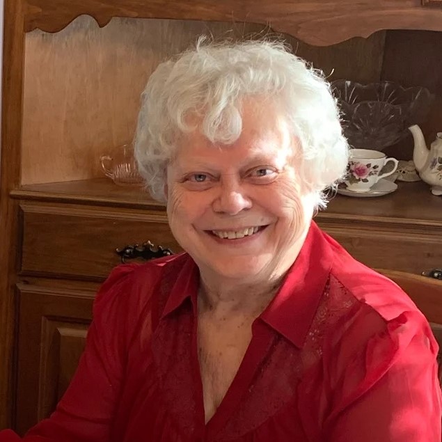 mature woman with white hair wearing red collared shirt smiling in front of wooden cabinet and white china tea set