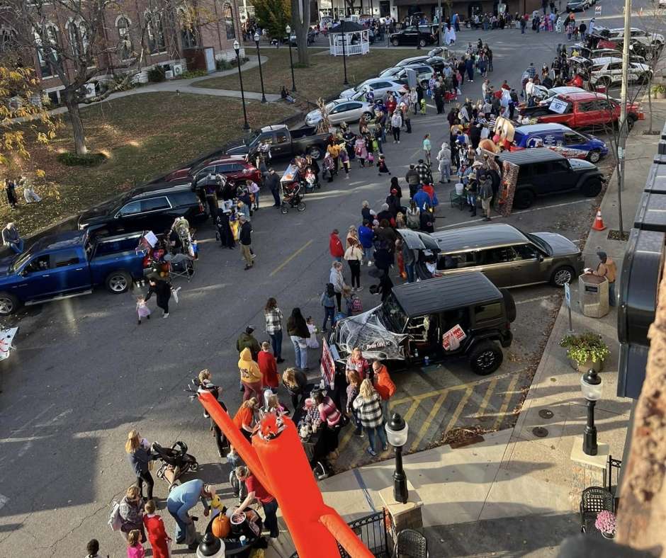 Aerial view of a lively outdoor event with numerous people gathering around parked cars. The scene is festive and bustling with vibrant autumn colors.