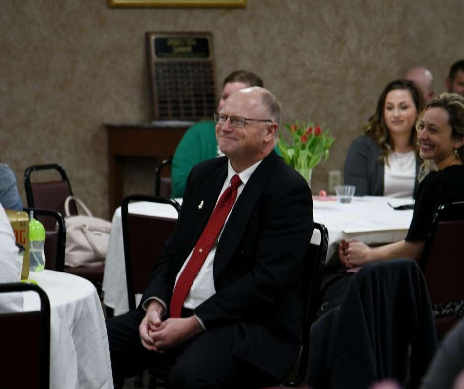 A man in a suit and red tie sits at a round table with others, smiling in a warmly lit room. Tulips and assorted drinks create a welcoming ambiance.
