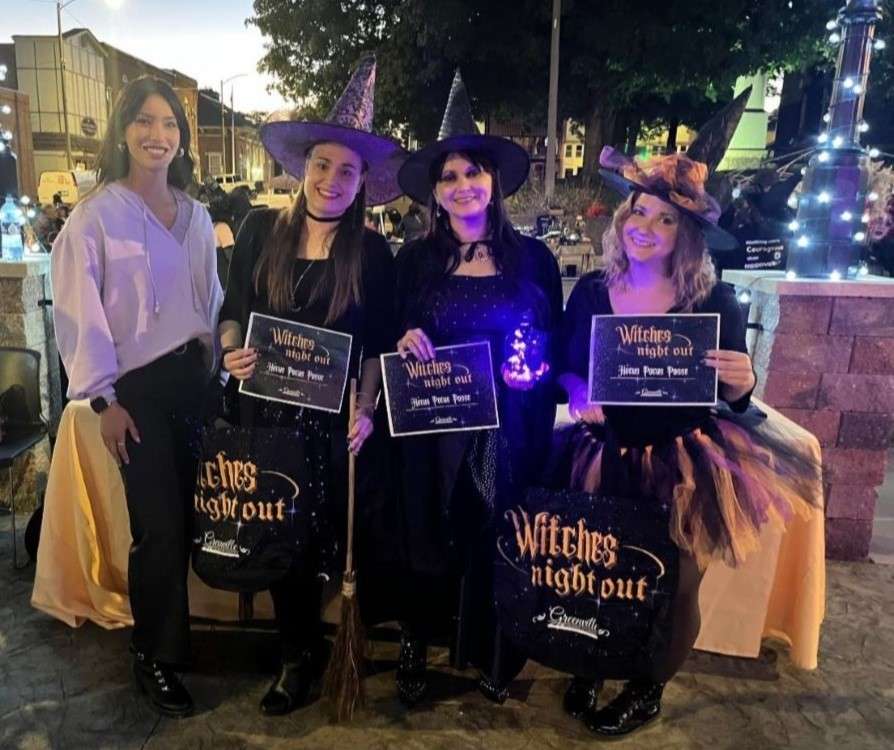 A group of four women outdoors, three dressed as witches with hats, holding Witches Night Out signs. The mood is festive and joyful.