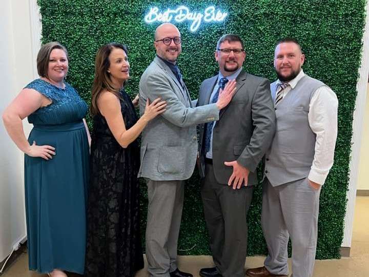 two ladies in formal dresses and three men in gray suits pose in a line standing in front of green wall and neon sign that reads Best Day Ever