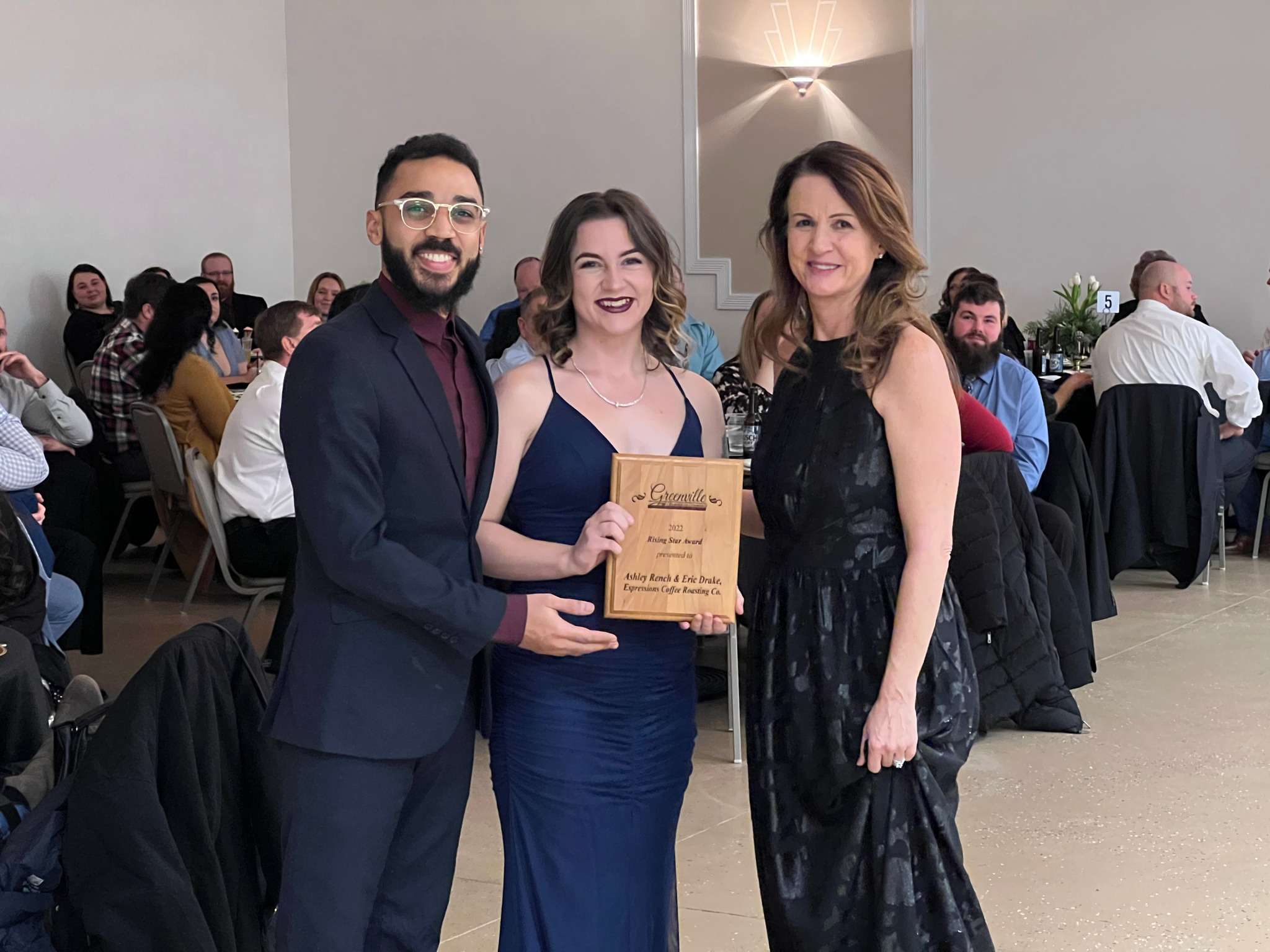 two ladies in formal dresses and one man in suit and glasses smile and hold a wooden plaque award. behind trio are event attendees seated at round tables.