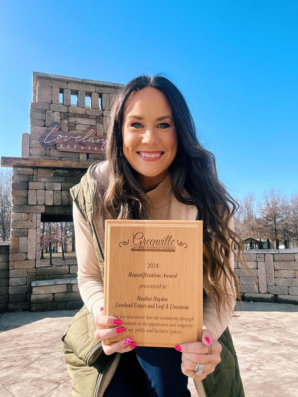 young woman outdoors in front of decorative stone facade, she holds wooden award plaque