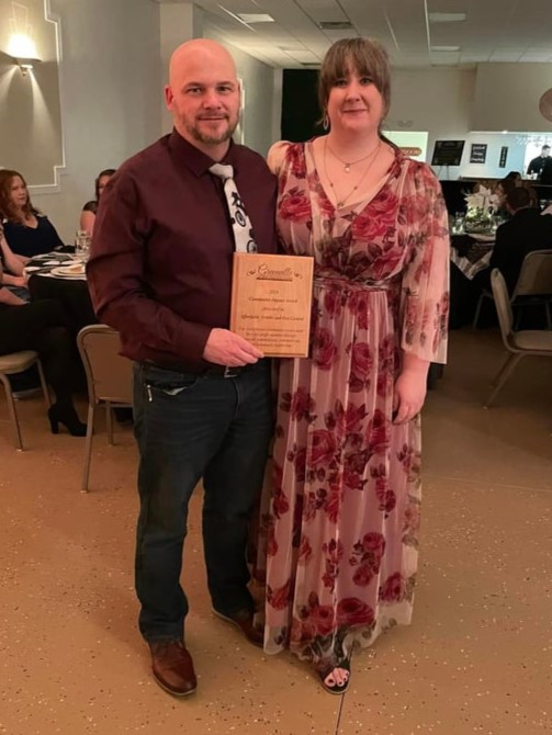 young bald man in dark red shirt, black trousers and tie and woman in long floral cocktail dress hold wooden award plaque