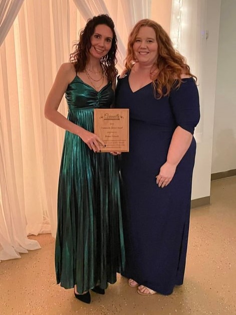 two women indoors in front of white sheer curtains, they hold wooden award plaque