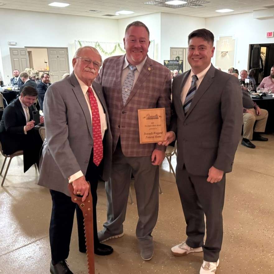 three men in gray suits patterened ties, one older man leans on a walking cane, they hold a wooden award plaque
