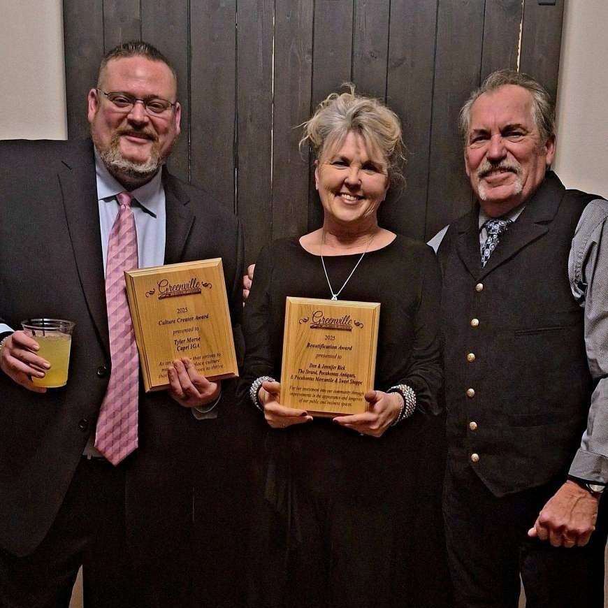 smiling middle age woman with updo hair, bling jewelry and long sleeved black dress flanked by two middle age men in ties and black suits, together they hold two wooden award plaques