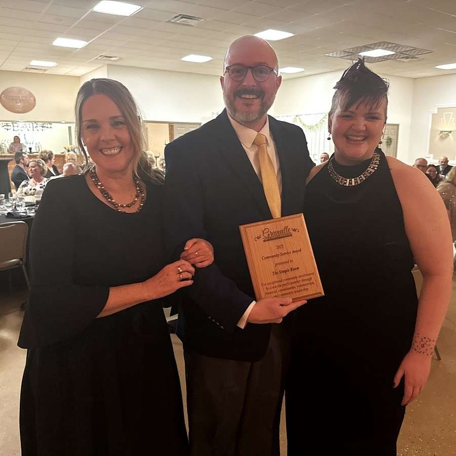 two women with large necklaces, one with purple spikey hair and a tattoo, both wear black formal dresses, between them is a young bald man in a suit who holds a wooden award plaque