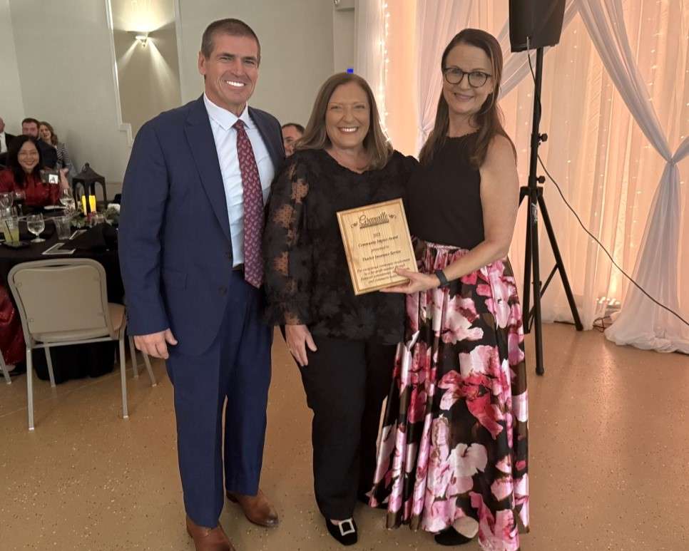 middle aged man in buzz cut hair and blue suit and red tie, and woman in black lacey shirt and dress trousers, woman with long dark hair wearing ankle length pink floral skirt holds wooden award plaque