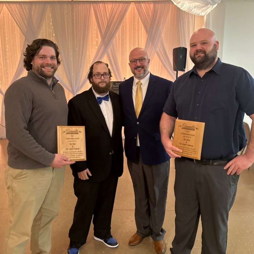 two young adult men in suits, two men in polo shirts and nice trousers, they hold two wooden award plaques