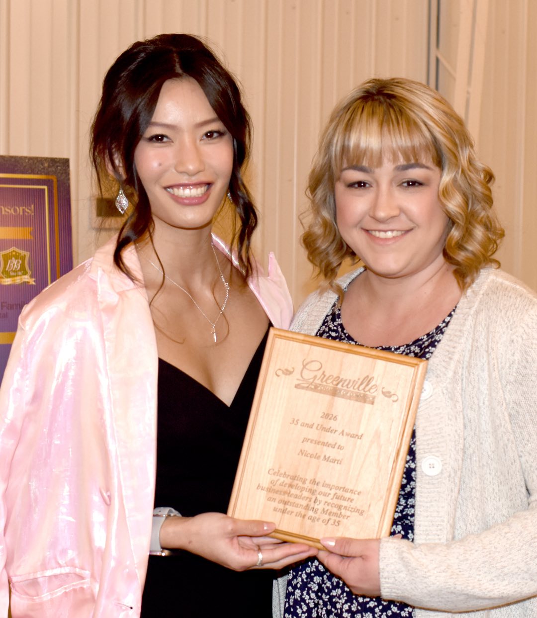 two women hold a wooden award plaque between them, one with dark hair and wearing a black dress under a pearlescent cape and the other with blond hair wearing a aptterend ddress under an ivory knit sweater