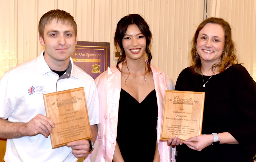 two young persons, a man in a white polo shirt with business logo and a woman in a black coctail dress, they stand on either side of a woman in a pearlescent cape and hold two wooden award plaques