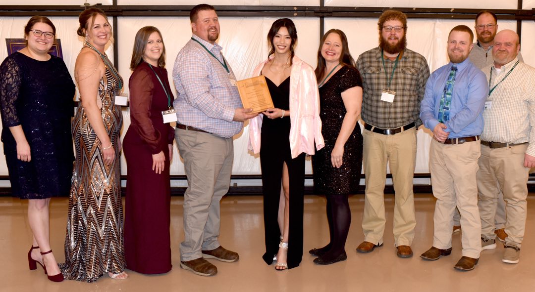 smiling group of five women in sparkling party dresses and five men in business casual attire, together they hold a wooden award plaque