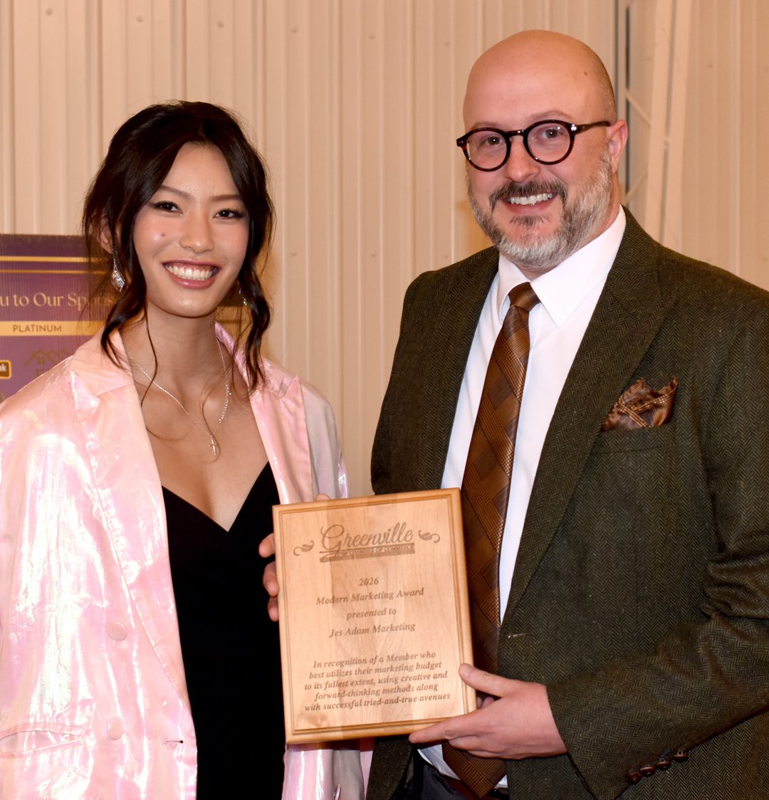 young woman on the left in black dress and pearlescent cape, bald man with salt and pepper beard on the right wearing a dark suit and tie, they hold a wooden award plaque