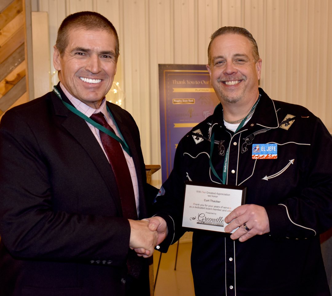 two men with graying short clipped hair, one wears a suit and tie, the other wears a Western style embroidered dress shirt