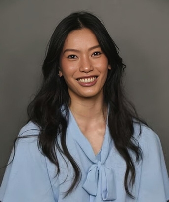 portrait headshot of a slim attractive woman in her twenties wearing a light blue blouse with attached tied scarf at V neckline. She has long black hair, almond shaped eyes, and wide smiling mouth.
